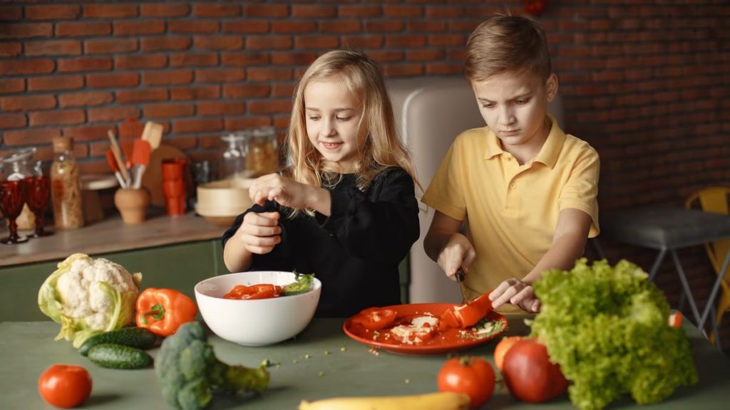 Serious boy cutting pepper for vegetable salad while standing near happy sister seasoning ingredients put into deep bowl in loft style kitchen